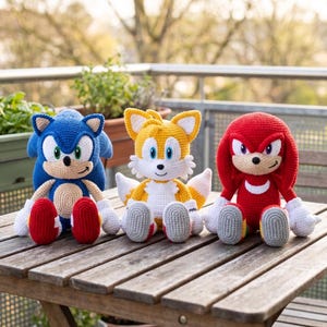 Op de afbeelding: Drie handgemaakte gehaakte knuffels die Sonic the Hedgehog, Tails en Knuckles voorstellen. De blauwe Sonic, gele Tails en rode Knuckles figuren hebben gedetailleerd stikwerk, witte accenten en geborduurde gelaatstrekken in een zittende houding.