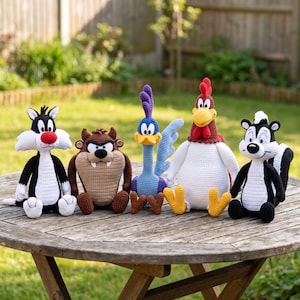 Op de afbeelding: Een verzameling van vijf gehaakte knuffelfiguren, waaronder een zwart-witte kat met een rode neus, een bruine Tasmaanse duivel, een blauwe en paarse roadrunner, een witte haan met een rode kam en een zwart-wit stinkdier.