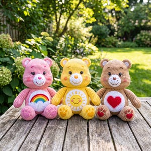 Könnte beinhalten: Drei gehäkelte Stoffbären sitzen in einer Reihe. Der linke Bär ist rosa mit einem Regenbogen-Bauchabzeichen. Der mittlere Bär ist gelb mit einem Sonnen-Bauchabzeichen. Der rechte Bär ist hellbraun mit einem roten Herz-Bauchabzeichen. Jeder Bär hat gestickte Gesichtszüge und herzförmige Pfoten.