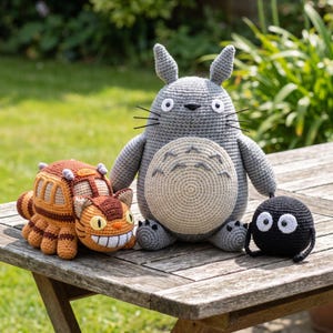 Pode incluir: Um conjunto de três peluches de croché, incluindo um Totoro grande cinzento com barriga creme, um autocarro em forma de gato castanho e laranja com várias pernas e janelas, e um pequeno espírito de fuligem preto com dois olhos brancos.