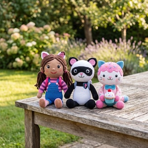 Könnte beinhalten: Drei handgefertigte Häkelpuppen sitzen nebeneinander. Die linke Puppe hat braune Haare, Katzenohren und eine blaue Latzhose. Die mittlere Puppe ist ein schwarz-weißer Panda mit Rucksack. Die rechte Puppe ist eine rosa Katze mit einem Cupcake.