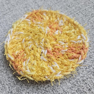 May include: A round, handmade crochet scrubby in shades of yellow, white, and peach. The scrubby has a textured, looped appearance, with visible yarn strands creating a fluffy, absorbent surface. The item is designed for cleaning.