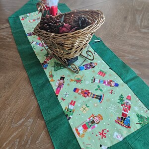 May include: A Christmas-themed table runner with a green border and a light green center featuring nutcracker and holiday graphics. A woven sleigh filled with pinecones and decorative items sits on the runner, with a small elf figurine in the background.