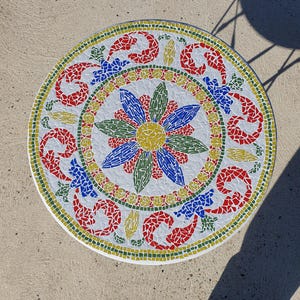 Op de afbeelding: Een rond tafelblad met een mozaïekontwerp met een centrale gele bloem omringd door blauwe en groene bloemblaadjes. De buitenrand toont rode, gele en groene geometrische patronen op een witte achtergrond, wat zorgt voor een levendig, kleurrijk oppervlak.