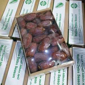 May include: A rectangular cardboard box filled with brown dried dates, wrapped in clear plastic film. The box is labelled with green text reading Madina Dates Season and features a palm leaf graphic, surrounded by several identical boxes.