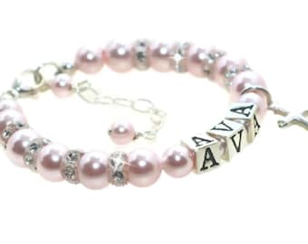 May include: A pink pearl bracelet with a silver charm and a silver cross. The bracelet has the name "AVA" spelled out in silver beads.