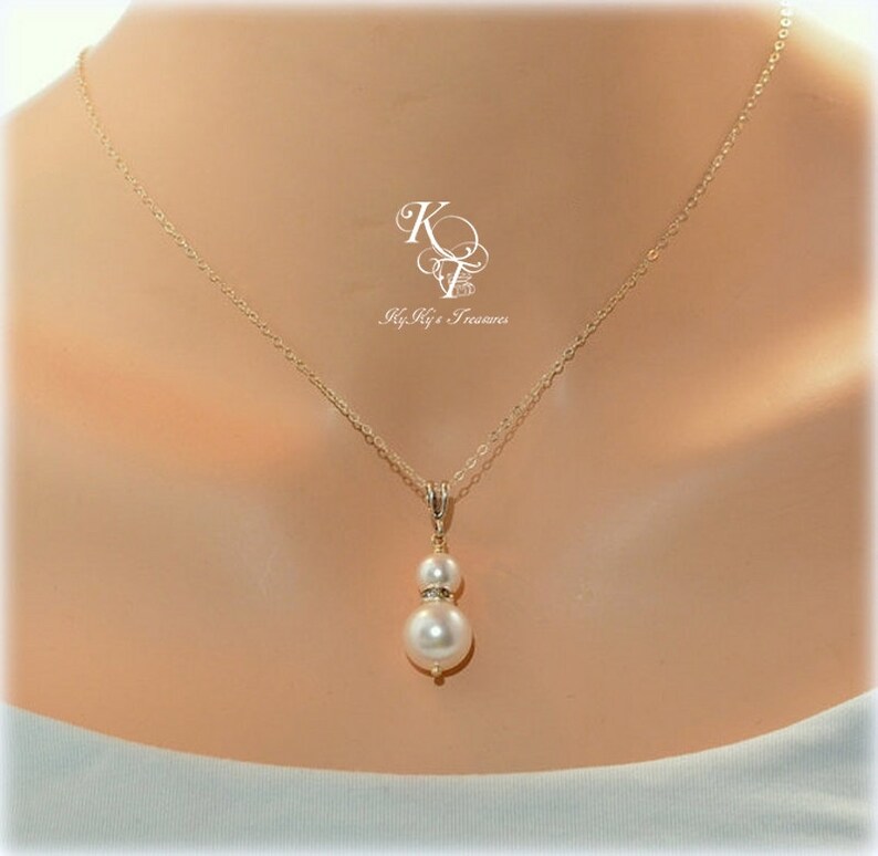 May include: A gold chain necklace with a double pearl pendant. The larger pearl is white and the smaller pearl is accented with small clear stones.