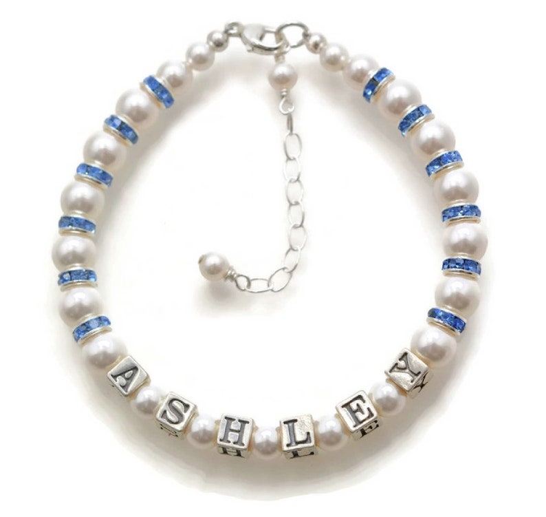 May include: A white pearl bracelet with blue crystal accents and a silver charm with the name "ASHLEY" spelled out.