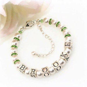 May include: A white pearl bracelet with green crystal accents and silver charm beads spelling out the name "Brooke".