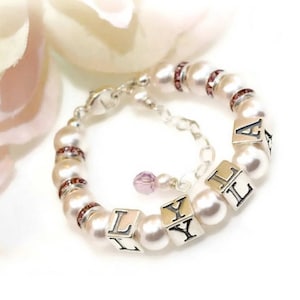May include: A white pearl bracelet with a silver clasp and pink crystal accents. The bracelet has silver letter beads that spell out the name "LYLA".