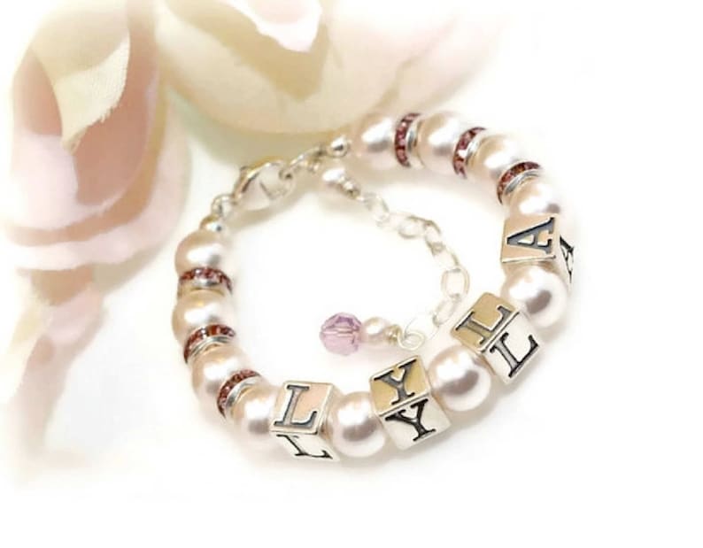 May include: A white pearl bracelet with a silver clasp and pink crystal accents. The bracelet has silver letter beads that spell out the name "LYLA".