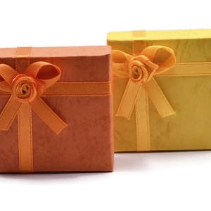 May include: Two square gift boxes, one in a burnt orange shade and the other in a pale yellow hue. Each box is wrapped with a matching satin ribbon that features a decorative rosette bow centered on the top lid of the box.
