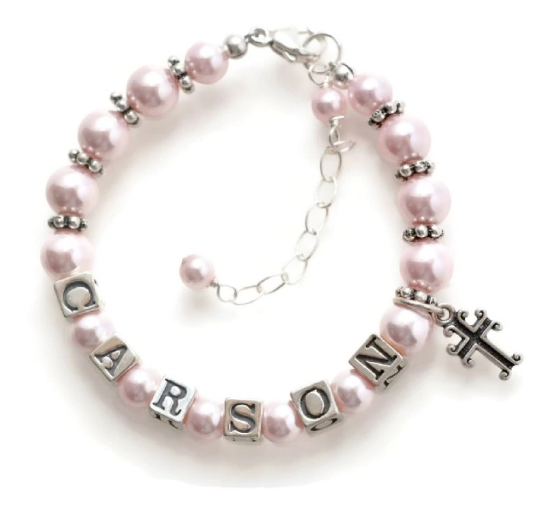 May include: A pink pearl bracelet with the name "CARSON" spelled out in silver lettered cubes. The bracelet has a silver cross charm and is adjustable. The pearls are approximately 0.3 inches in diameter.