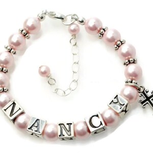 May include: A pink pearl bracelet with the name "NANCY" spelled out in silver lettered blocks. The bracelet has a silver cross charm and silver accents between the pearls. The bracelet is adjustable.