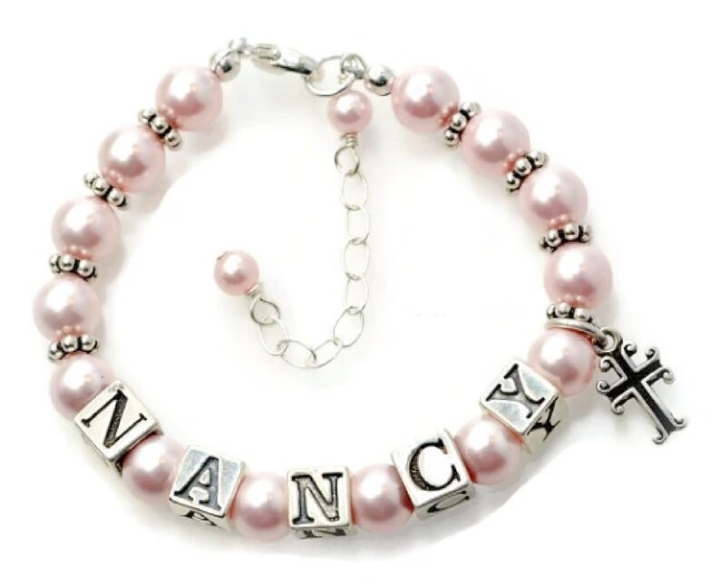 May include: A pink pearl bracelet with the name "NANCY" spelled out in silver lettered blocks. The bracelet has a silver cross charm and silver accents between the pearls. The bracelet is adjustable.