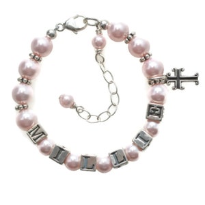 May include: A pink pearl bracelet with silver accents and the name "MILLIE" spelled out in silver letter beads. A silver cross charm hangs from the bracelet, which has an adjustable chain closure.