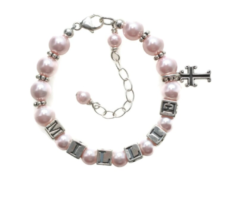 May include: A pink pearl bracelet with silver accents and the name "MILLIE" spelled out in silver letter beads. A silver cross charm hangs from the bracelet, which has an adjustable chain closure.