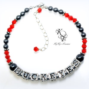 May include: A beaded bracelet featuring the word "BUCKEYES" spelled out in silver letter beads. The bracelet includes black and red beads, with a silver chain for adjustable sizing. The bracelet is likely a fan item.