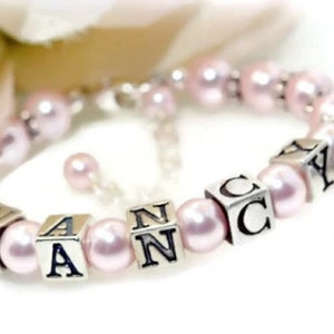 May include: A delicate bracelet featuring pale pink pearls and silver lettered cubes spelling "NANCY". A silver cross charm dangles from the bracelet, which is strung on a silver chain.