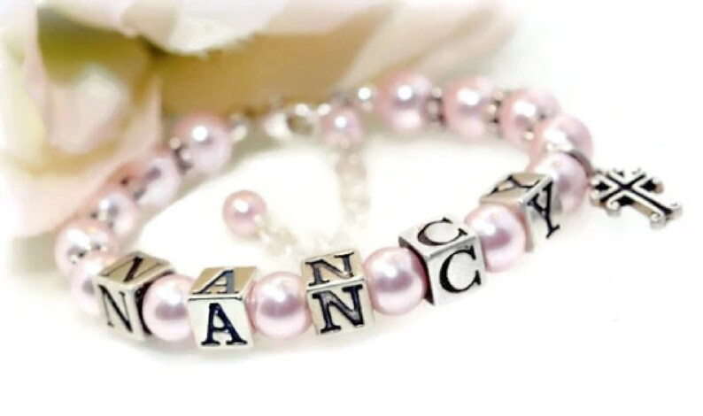May include: A delicate bracelet featuring pale pink pearls and silver lettered cubes spelling "NANCY". A silver cross charm dangles from the bracelet, which is strung on a silver chain.