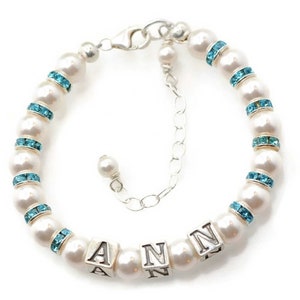 May include: A white pearl bracelet with a silver clasp and blue crystal accents. The bracelet has a charm with the name "ANN" spelled out in silver.