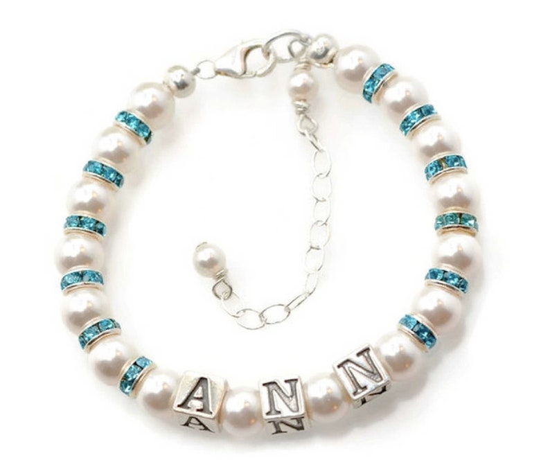 May include: A white pearl bracelet with a silver clasp and blue crystal accents. The bracelet has a charm with the name "ANN" spelled out in silver.