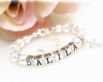 May include: A silver bracelet with white pearls and a charm with the name "DALILA" spelled out in black letters. The bracelet also has a small silver cross charm.