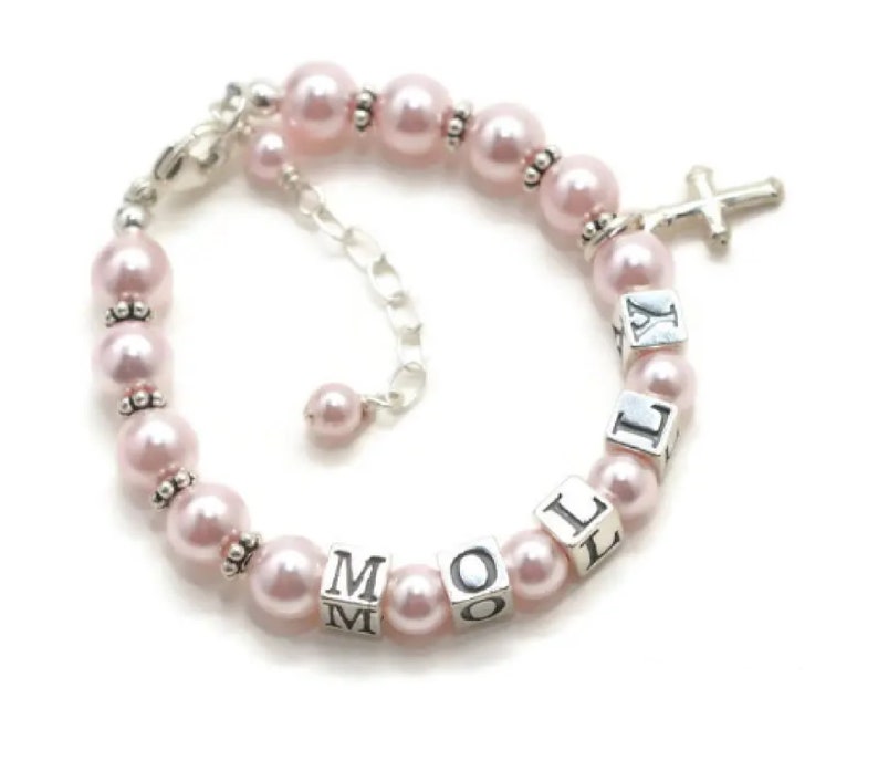 May include: A pink pearl bracelet with silver accents and a cross charm. The bracelet features the name "Mollie" spelled out with silver letter beads. The bracelet is adjustable.