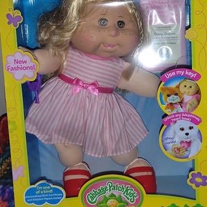 May include: A Cabbage Patch Kids doll with blonde hair, freckles, and a pink and white striped dress with a pink bow. The doll wears red shoes and holds a purple key. The packaging includes text such as New Fashions, Use my key, and Penny Luciana.