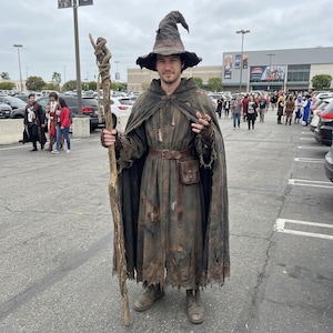 May include: A dark brown, distressed wizard costume featuring a tattered, floor-length hooded robe with a leather belt and small pouch, a matching pointed hat with a wide brim, and a tall, gnarled wooden staff wrapped in twine.