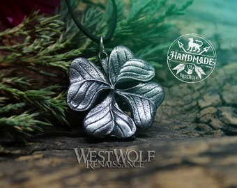 May include: A silver four-leaf clover pendant necklace on a black cord, resting on a wooden surface with green foliage. The pendant is detailed with textured lines and the words "Westwolf Renaissance" are visible below it. A circular logo with the words "Handmade Westwolf" is visible in the upper right corner.