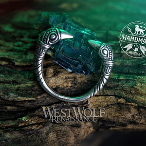 May include: A silver ring with two bird heads on the sides. The ring is on a wooden surface with a green background.