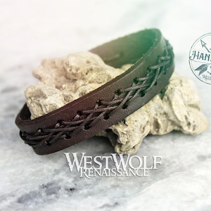 May include: A brown leather bracelet with a braided design. The bracelet is handmade and has a small leather tag with the words "Handmade Westwolf" on it.