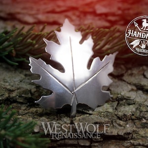 Puede incluir: Un colgante de hoja de arce de plata con un diseño detallado. El colgante está hecho a mano y presenta un fondo rústico.