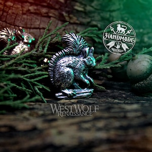 May include: A silver squirrel figurine with a bushy tail, sitting on a wooden surface with green foliage and a brown acorn. The figurine is handmade and has a rustic, vintage look.