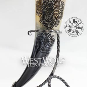 May include: A handcrafted drinking horn with intricate Celtic knotwork and a decorative stand. The horn is a light tan color with dark etched designs. The stand is made of wrought iron. The text "WESTWOLF RENAISSANCE" is visible.