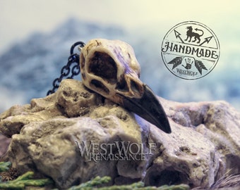 May include: A raven skull pendant with a black chain, resting on a textured stone. The skull is off-white with a dark beak. The image includes a "HANDMADE" logo and the text "WESTWOLF RENAISSANCE".