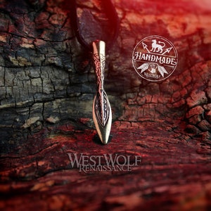 May include: A bronze spearhead pendant with intricate details, hanging on a black leather cord. The pendant is on a rustic wooden surface with a red-toned background. The image includes the text "Handmade by Westwolf".