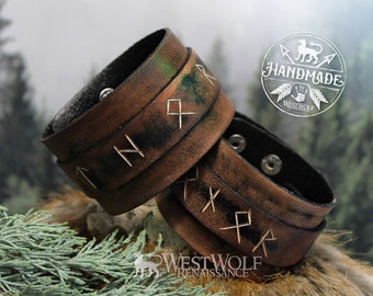 May include: Two brown leather bracelets with silver-coloured runes engraved on them. The bracelets are worn and have a distressed look. The bracelets are on a brown surface with green foliage in the background.
