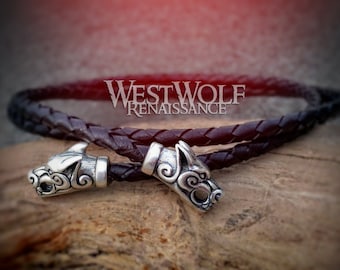 Peut inclure: Un bracelet en cuir tressé noir avec deux pendentifs en argent en forme de tête de loup. Le bracelet est posé sur une surface en bois. Westwolf Renaissance.
