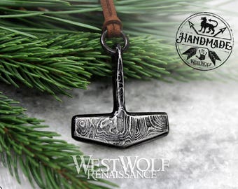 Puede incluir: Un collar con un colgante de Mjolnir de color plateado con un cordón de cuero marrón. El colgante tiene una superficie detallada y texturizada y está hecho a mano. El colgante está sobre un fondo blanco con una rama de pino verde en el fondo.