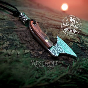 May include: A handmade, silver and wood,  knife pendant with a  patterned blade and a wooden handle. The pendant is hanging from a black leather cord with a silver bead. The pendant is on a wooden surface with the text "Handmade" and "Westwolf Renaissance" visible.