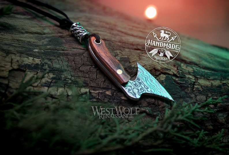 May include: A handmade, silver and wood,  knife pendant with a  patterned blade and a wooden handle. The pendant is hanging from a black leather cord with a silver bead. The pendant is on a wooden surface with the text "Handmade" and "Westwolf Renaissance" visible.