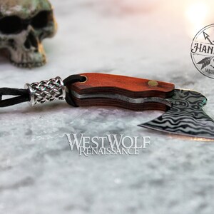 May include: A small, black leather cord with a silver braided bead attached to a miniature axe with a wooden handle. The axe blade is made of stainless steel with a wavy pattern. The axe is handmade and has the words "Handmade Westwolf" on the handle.