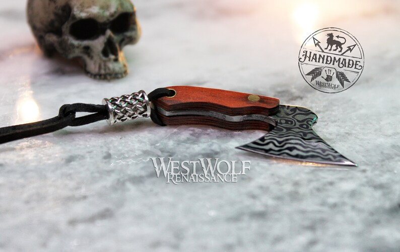 May include: A small, black leather cord with a silver braided bead attached to a miniature axe with a wooden handle. The axe blade is made of stainless steel with a wavy pattern. The axe is handmade and has the words "Handmade Westwolf" on the handle.