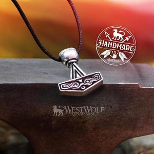 Könnte beinhalten: Eine silberfarbene Halskette mit Anhänger, die eine detaillierte Nachbildung von Thors Hammer, Mjolnir, zeigt. Der Anhänger hängt an einem schwarzen Band. Das Bild enthält den Text "HANDMADE" und "WestWolf Renaissance". Der Anhänger liegt auf einer dunklen Metalloberfläche.