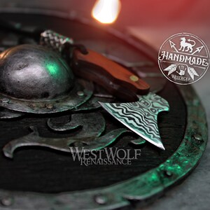 May include: A small, handmade axe with a wavy, black and white patterned blade. The axe is resting on a dark, circular shield with a green and silver design. The shield has the text "Westwolf Renaissance" on it.