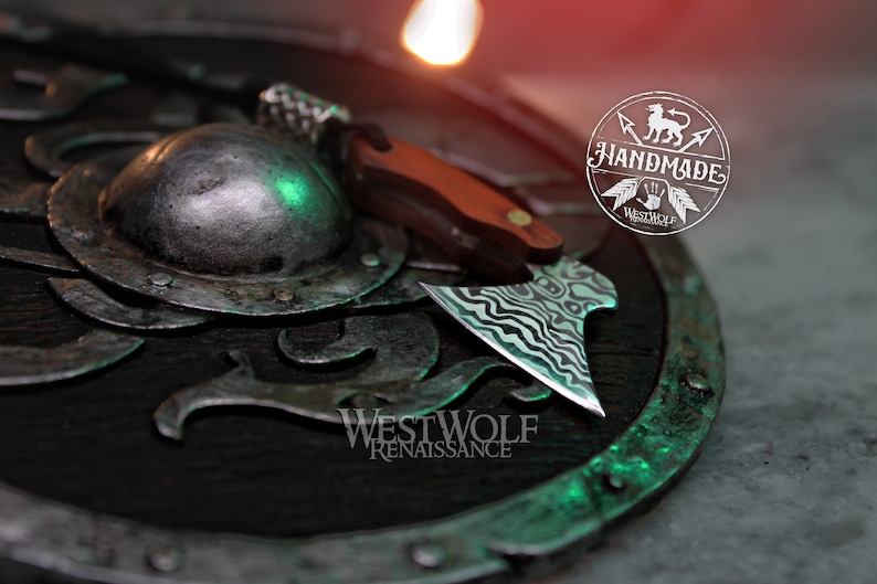 May include: A small, handmade axe with a wavy, black and white patterned blade. The axe is resting on a dark, circular shield with a green and silver design. The shield has the text "Westwolf Renaissance" on it.