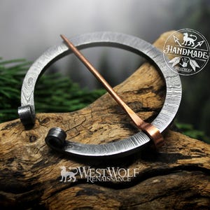 Puede incluir: Broche plateado hecho a mano con un alfiler de color cobre, sobre un fondo de madera rústica. El broche tiene un diseño circular con una textura estampada. La imagen incluye el texto "HANDMADE" y "WESTWOLF RENAISSANCE".