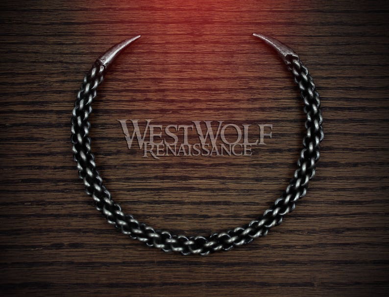 May include: A silver chain necklace with two pointed ends, forming a circle. The chain is made of linked rings, creating a textured look. The necklace is on a dark brown wooden surface.